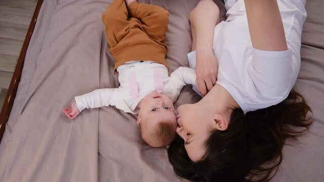 Top view of cute mum and baby daughter lying on cozy bed have fun use cell phone watch cartoons, mom with infant girl making online call vlog. Parental control, modern tech, younger generation concept