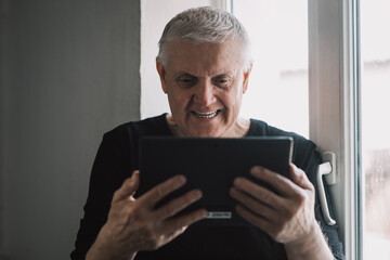 a close-up portrait of a cheerful, cheerful old man holding an electronic tablet with high-speed internet in his hands in a bright room by the window
