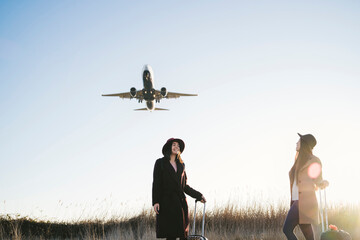 Friends waiting with wheeled luggage on roadside, airplane flying above