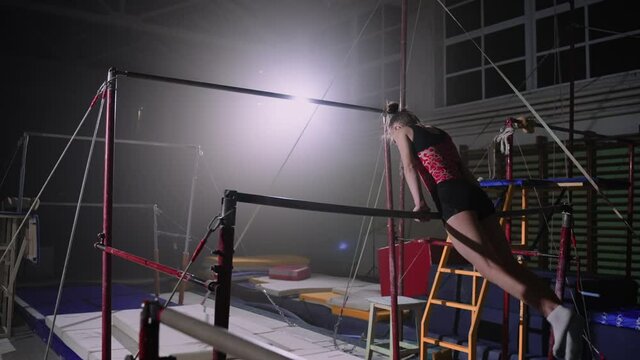 sporty female gymnast is performing exercises on uneven bars in gymnastic hall at evening, female artistic gymnastics