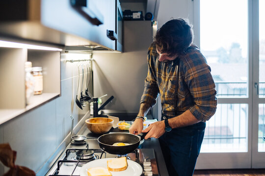 Mid adult man preparing fried eggs at kitchen hob while making smartphone call