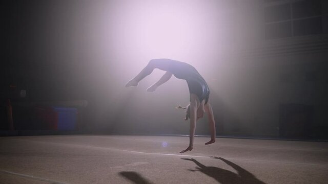 sporty adolescent girl is doing two somersaults back on spring floor, training in gymnastic hall alone, artistic gymnastics