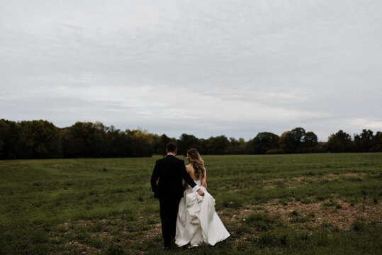 Romantic young groom carrying bride's wedding dress train in field, rear view