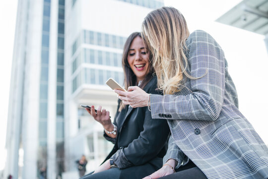 Two young businesswomen sitting outside office building looking at smartphones, Turin, Piemonte, Italy