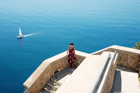Young woman enjoying sunny day by sea, sailboat in background, Alghero, Sardegna, Italy
