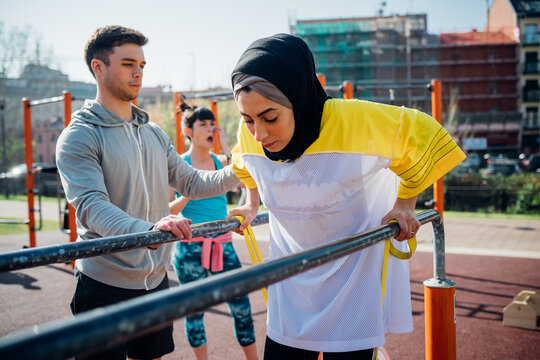 Calisthenics class at outdoor gym, male trainer encouraging young woman on parallel bars