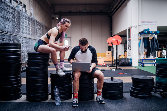 Young woman and man training together in gym, sitting on weights looking at smartphone and laptop