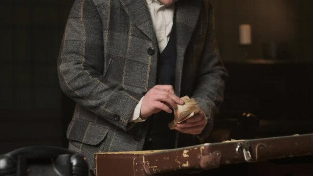 Mafia gangster in plaid suit gets his loot in form of old money from suitcase of brown leather. Criminals accountant counts money.
