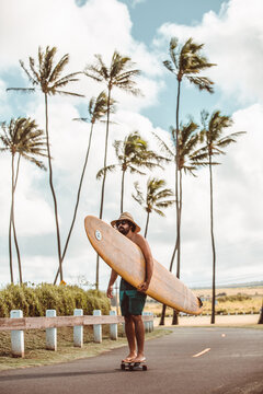 Mid adult male skateboarder carrying surfboard, skateboarding on coastal road, Haiku, Hawaii, USA