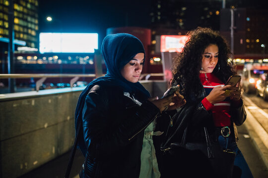 Young woman in hijab with friend looking at smartphones in city at night