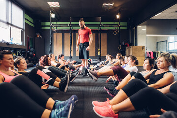 Large group of women training in gym with male trainer, sitting on floor with legs raised