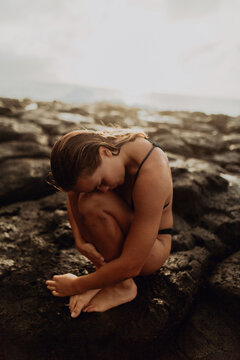 Woman sitting curled up on rocks by sea, Princeville, Hawaii, US