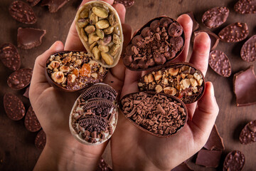 Top view of hands holding six mini stuffed easter eggs on the foreground. Lots of mini easter eggs and cracked chocolates on a wooden table at the background. 