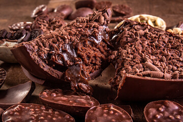 Close up of a cracked stuffed chocolate easter egg with grated chocolate on the top and lots of mini easter eggs and chocolates all around it on a wooden table.