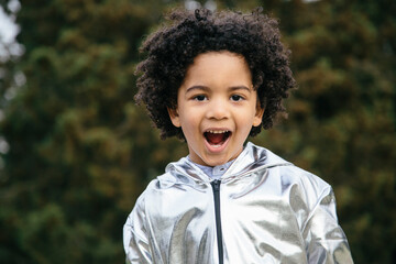 Black boy wearing a silver clothing, looking at the camera, screaming and smiling. In a park...