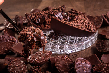 Spoonful of a cracked stuffed chocolate easter egg with grated chocolate on the top and lots of mini easter eggs and chocolates all around it on a glass stand on a wooden table.