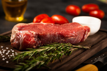 Fresh raw steak on wooden board on a dark background with salt, tomatoes and rosemary.
