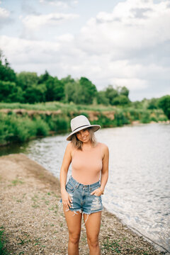 Young blonde woman wearing shorts and hat standing on a riverbank, smiling at camera.