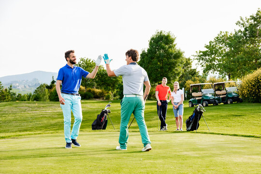 Friends giving high five on golf course, couple watching in background