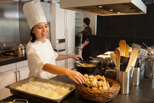Female chef wearing chef's hat standing at worktop in commercial kitchen, reaching for piece of ginger.