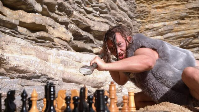 Actor playing caveman with painted face in fur vest threatens with stone spear to pieces on chessboard at rock slope on sunny day