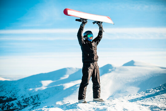 A person wearing a black ski suit, helmet and goggles on top of a mountain holding a red snowboard above their head.