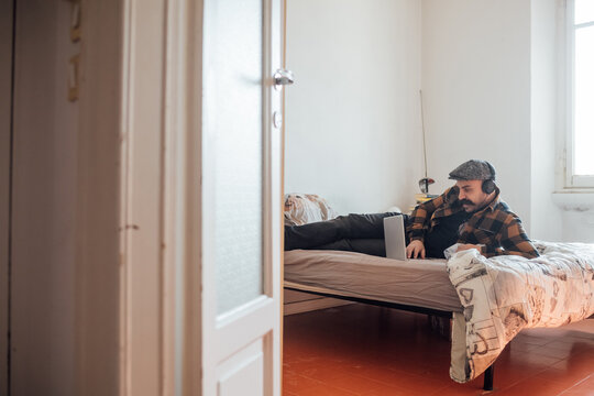 Man With Big Moustache Wearing Flat Cap And Headphones Lying On Bed With Laptop Computer During Corona Virus Crisis.