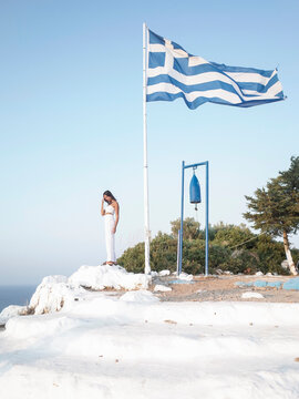 Woman standing next to Greek flag on rocky whitewashed vantage point on a Greek island.