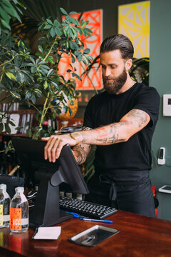 Bearded, tattooed bartender standing at counter, using computerized till.