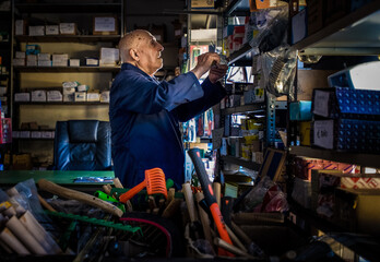 An ironmonger, elderly man in a boiler suit searching through shelves stacked with useful household items and tools. 