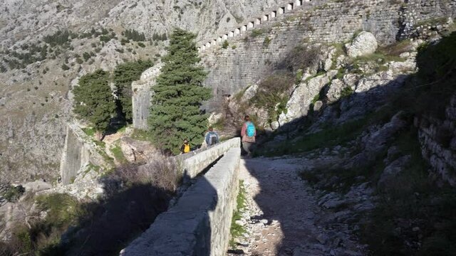 Tourists with backpacks climb down along rocky path with stone parapet at old Kotor fortress on sunny spring day backside view