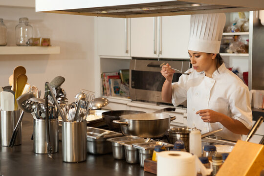Female chef wearing chef's hat standing at stove in a kitchen, tasting food.