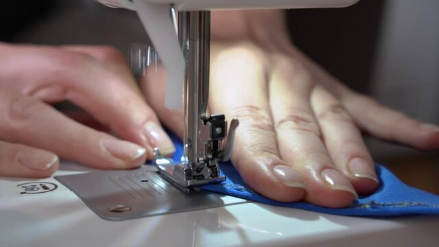 Skilled seamstress overcasts or housewife edge of blue serge fabric with contemporary sewing machine at workplace in room extreme closeup