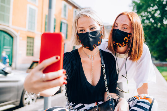 Two young women wearing face masks during Corona virus, sitting on a riverbank, taking selfie. 
