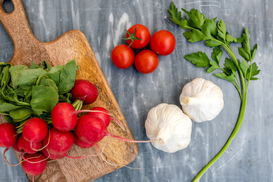 High angle close up of freshly picked radishes, garlic and flat leaf parsley and wooden chopping board on grey marble.