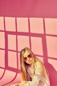 Portrait of girl with long blond hair wearing sunglasses and jacket, on pink background.