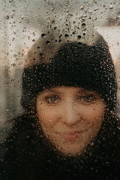 Head and shoulders portrait of woman wearing knit hat, looking at camera through window covered in raindrops.