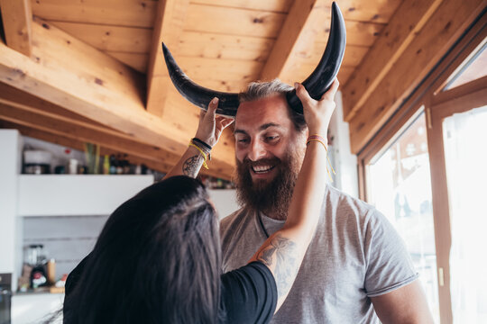 Woman Holding Cow Horns Onto Head Of Smiling Bearded Man With Brunette Hai.