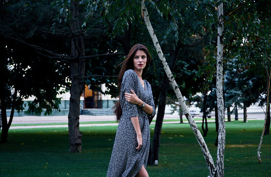 Portrait of woman with long brown hair, wearing long patterned wrap dress, standing in a park. 