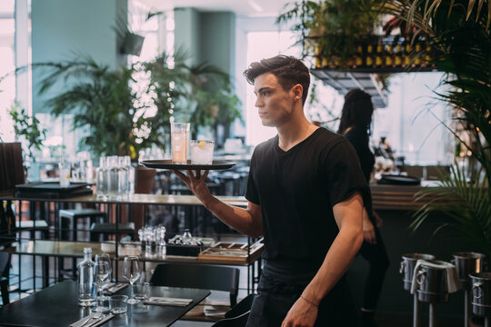 Young man wearing black clothes working in bar, carrying drinks on a tray.