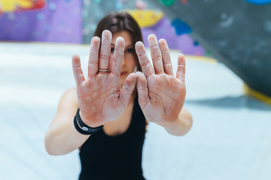 Woman In Black Outfit Standing In Front Of Indoor Rock Climbing Wall, Holding Out Chalk Powdered Hands.