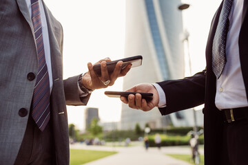 Two businessmen wearing suits standing outdoors, checking their mobile phones.