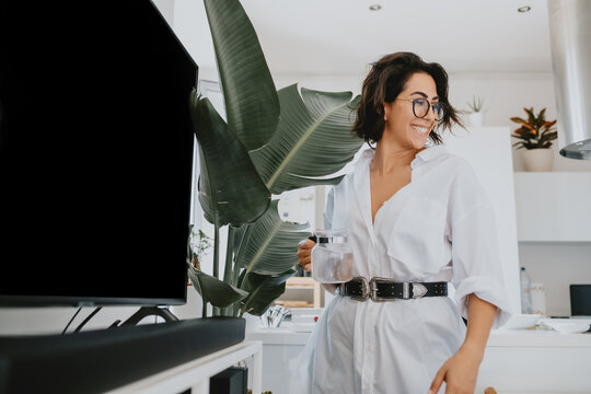 Smiling Woman With Brown Hair Wearing Glasses Standing In An Apartment, Watering Plant.