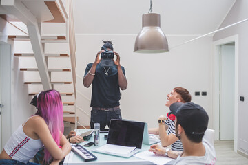 Group of young men and women gathered around a table, using laptops, tablets and virtual reality headset.
