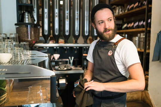 Bearded barista wearing apron working behind counter in a cafe.