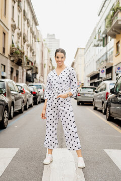 Portrait of smiling woman wearing white and black polka dot jumpsuit, standing on pedestrian crossing.