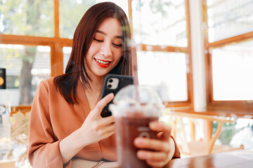 Smiling beautiful young Asian female food blogger holding a take away glass of shake and smoothie while taking picture on phone