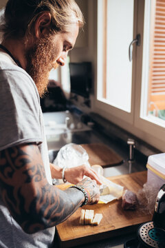 Bearded tattooed man with long brunette hair standing in a kitchen, preparing food.
