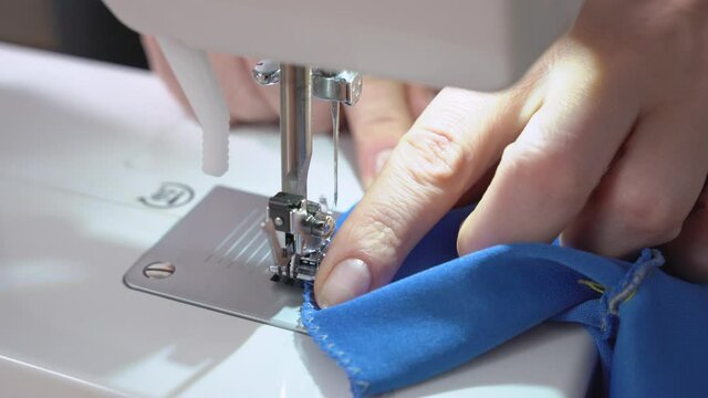 Skilled seamstress overcasts edge of blue serge fabric with contemporary sewing machine at workplace in room extreme closeup