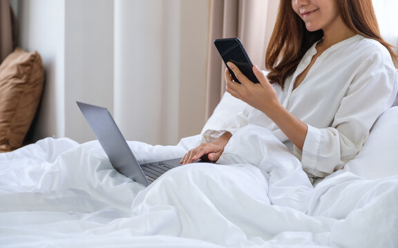 Closeup image of a beautiful young asian woman using mobile phone and laptop computer while lying down on bed